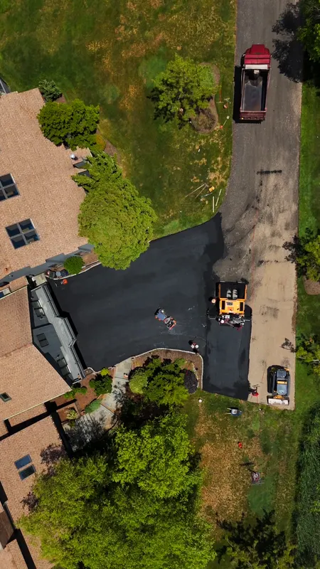 Overhead view of a residential driveway being paved beside the house, with fresh asphalt and the crew at work