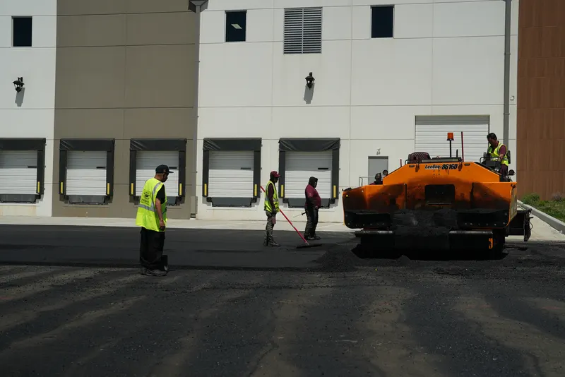 Paving machine laying asphalt along a warehouse while the crew works the edges
