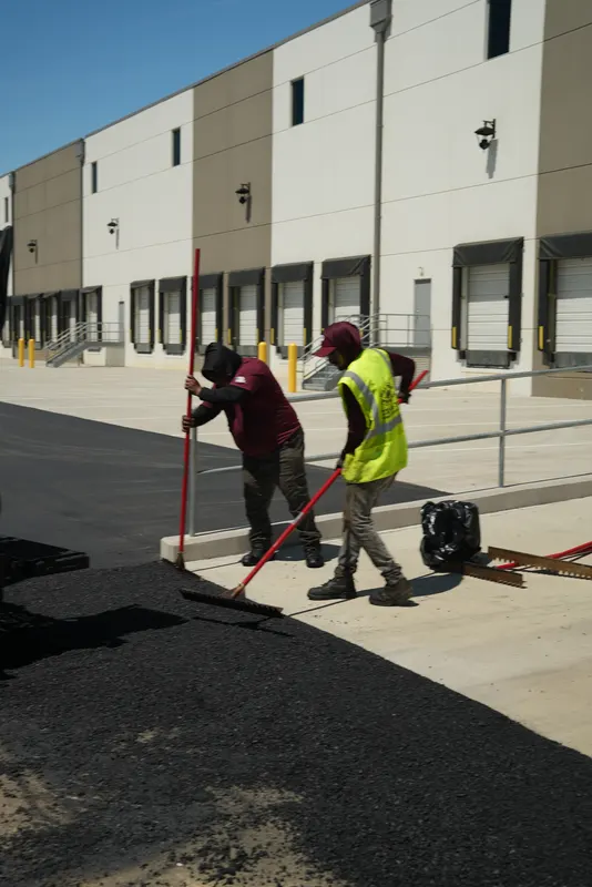 Crew raking and finishing the asphalt edge along a curb at a warehouse lot