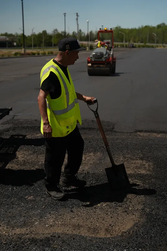Crew member working hot asphalt with a shovel while the roller compacts behind him