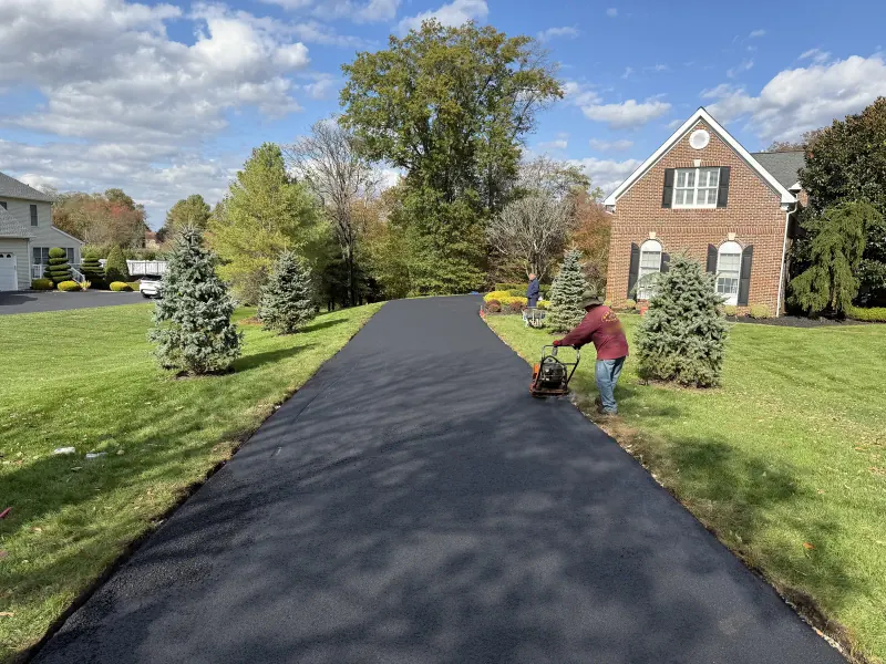 Crew compacting the edges of a new asphalt driveway in front of a brick home in the fall