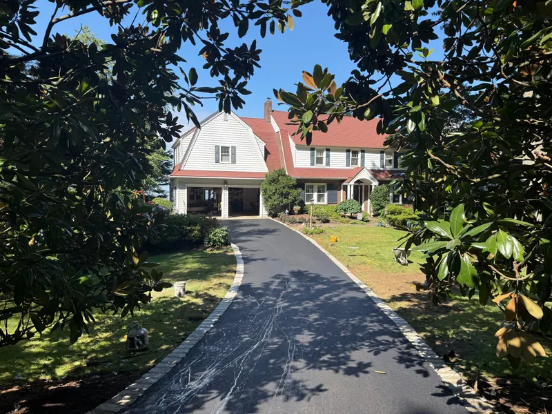 Long new black asphalt driveway with cobblestone edging leading up to a large colonial home, framed by magnolia trees