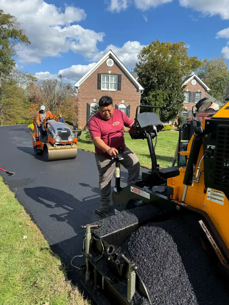 Crew member running the paving machine on a residential driveway while a roller compacts the new asphalt