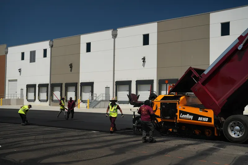 Paving crew, paving machine and dump truck working across a commercial parking lot